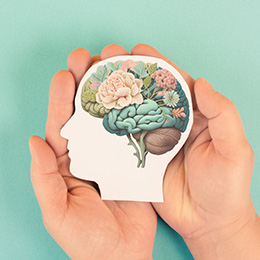 Hands holding a cardboard cut out of a human face and brain, with the brain having flowers, leaves on it symbolising mental health.